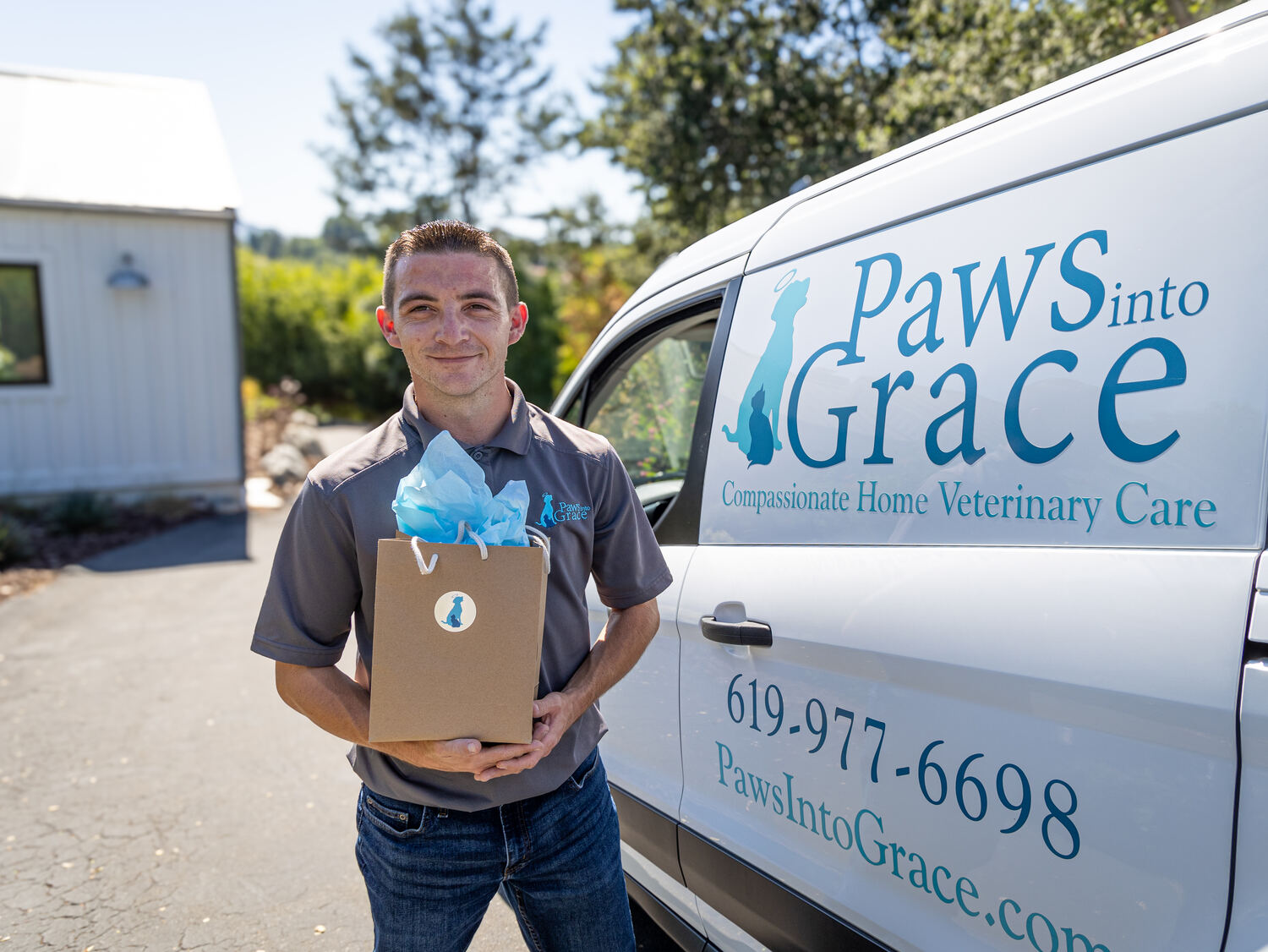 A man standing outside a van, holding a gift bag with light blue tissue paper in front of the 'Paws into Grace' logo.