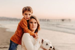 A woman sits on the beach holding a white dog while a young boy hugs her from behind