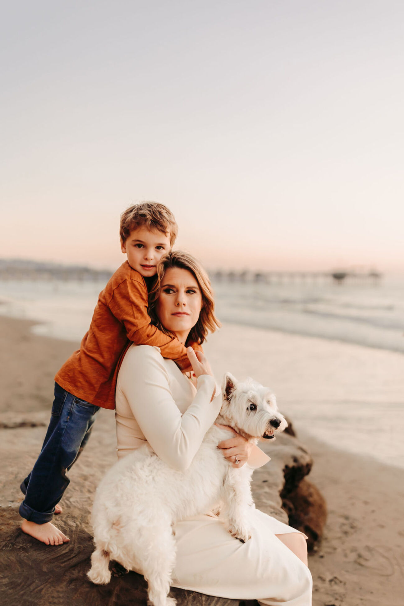 A woman sits on the beach holding a white dog while a young boy hugs her from behind
