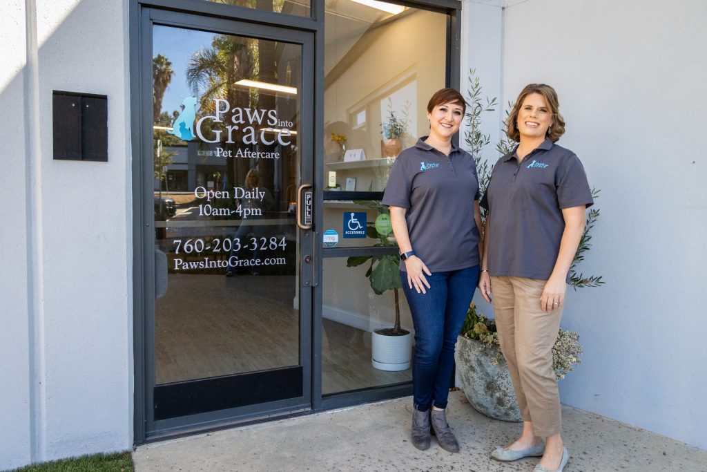 Two women stand outside the "Paws into Grace" Pet Aftercare business, smiling in matching shirts