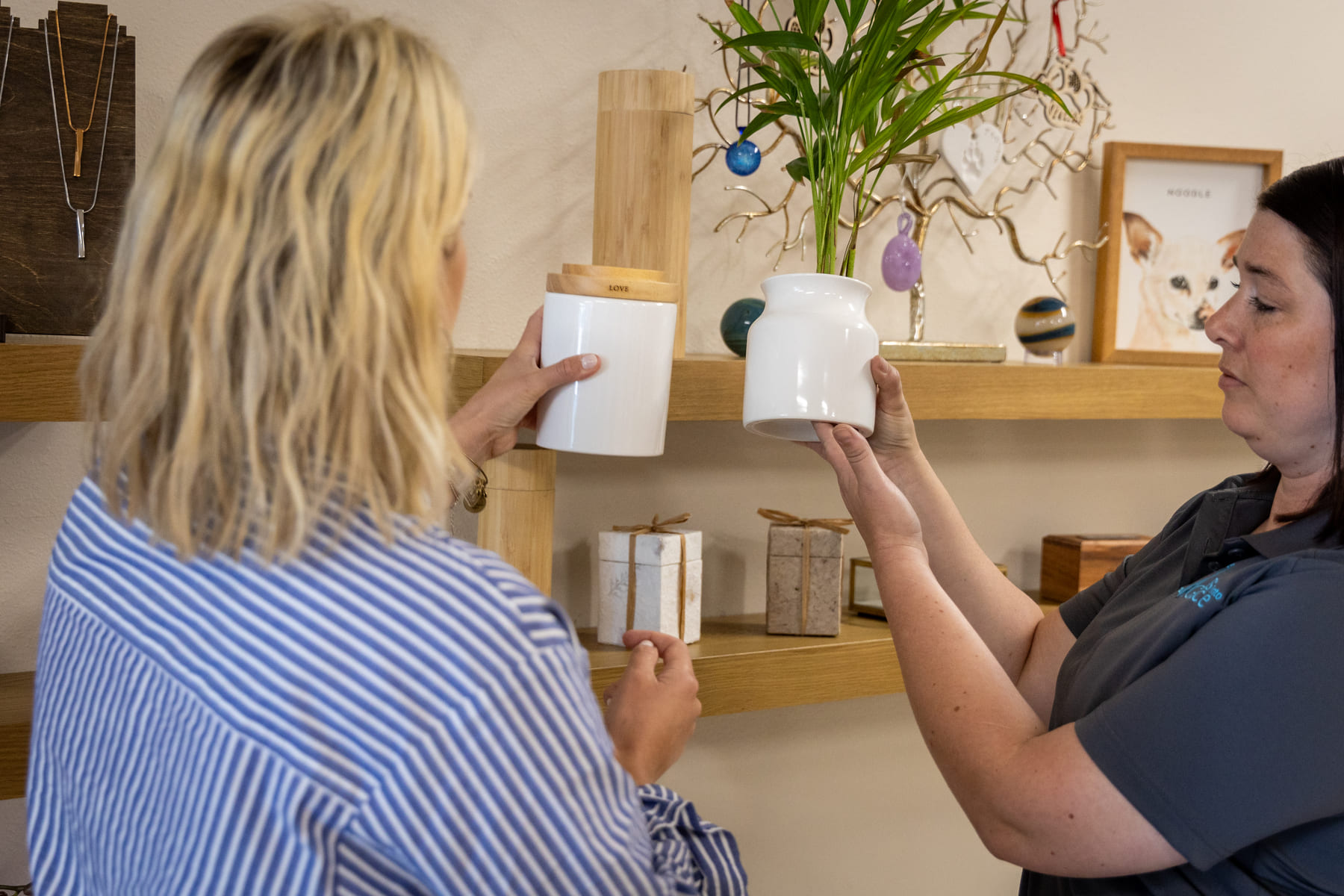 Two women examine white urns in a memorial display area with keepsakes